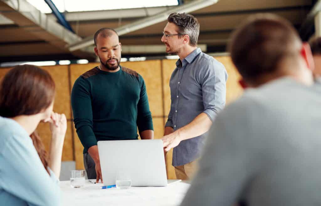 Zwei Kolleg*innen diskutieren über ihre Arbeit, während sie an einem Schreibtisch mit einem Laptop stehen.